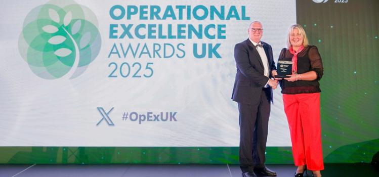 Woman in bright red trousers accepting an award trophy from a besuited man at an awards ceremony. Background presentation screen reads 'Operational Excellence Awards UK 2025 / #OpExUK'.
